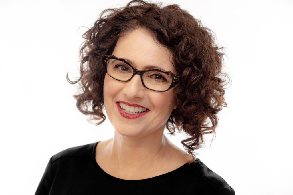 A portrait of a smiling woman, (Ayala Fader), with wavy medium length hair and glasses.