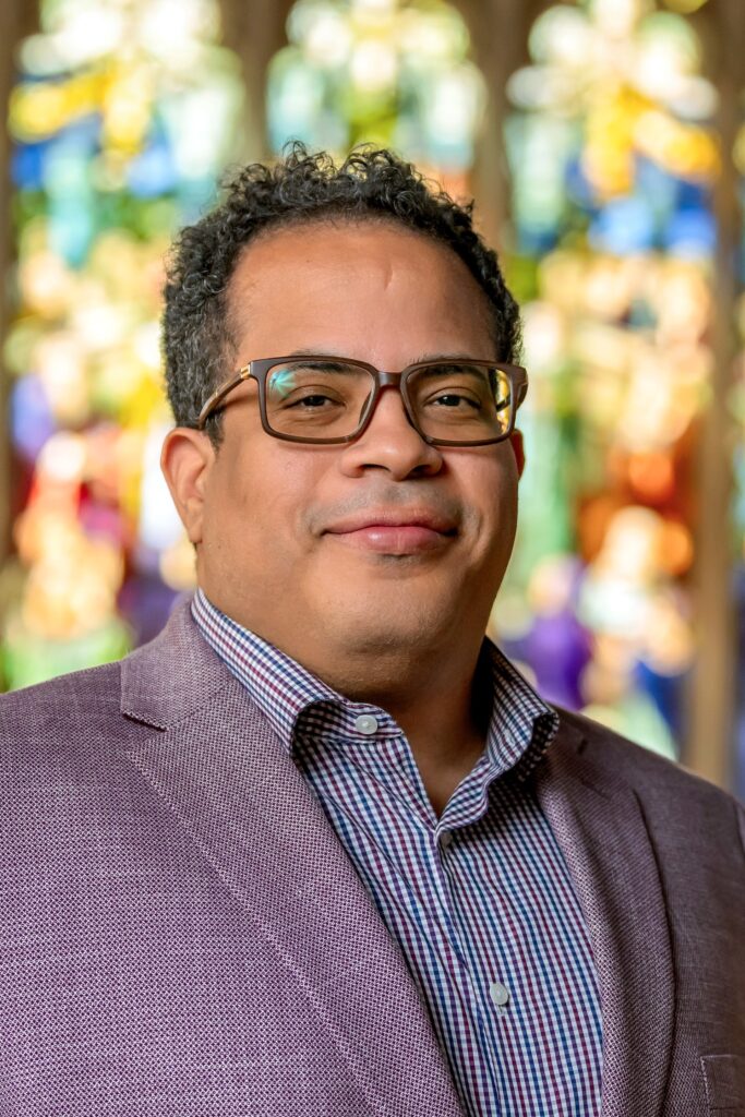 An interior portrait photo of a smiling man with short hair, wearing glasses, and open collar button down shirt and a blazer jacket, posing in front of stained glass window.